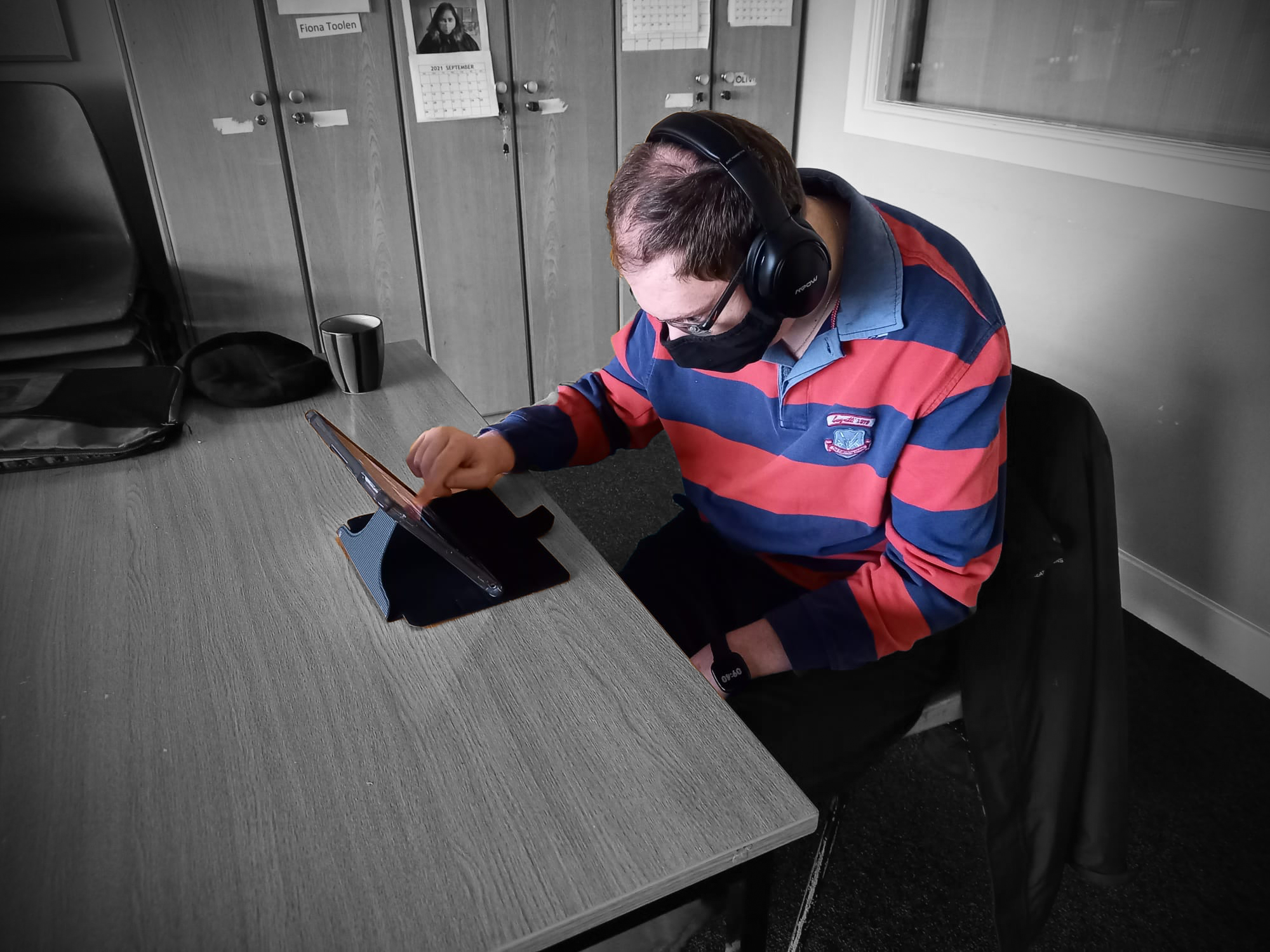 This is a photograph of one of the Sound On! Artists. He is sitting down at a table listening to sounds that he recorded using Whats App on his tablet for the Sound On! project. He is a white man in his thirties. He is wearing black headphones, black glasses a blue and red striped top with a collar, and a black face mask. There are closed presses and stacked chairs in the background. The colour of the background in the photograph has been altered to black and white.