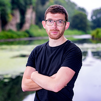A circular photograph of Artist AlanJames Burns, one of the lead artists on the Sound On! project. A white man in his thirties with brown hair, a red beard and glasses. He is wearing a black t-shirt and he has his arms folded. There are trees and a lake out of focus in the background.
